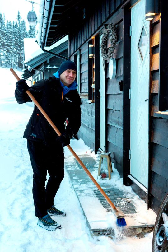Mann fjerner snø med kost utenfor hytte